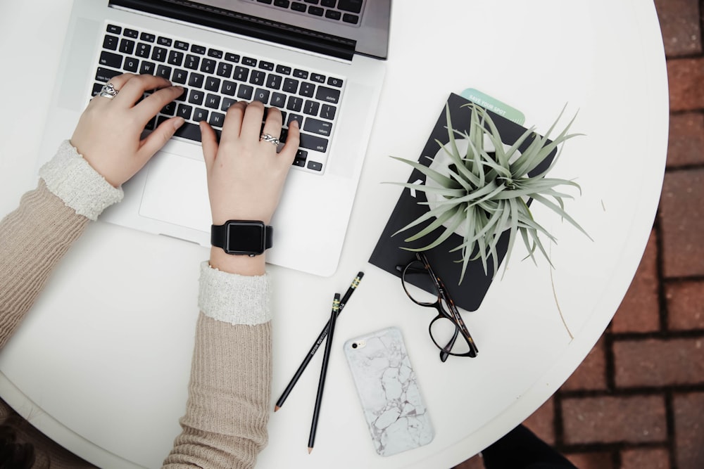 A person typing on a netbook with plants, pencils, a smartphone and glasses on the desk table