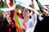 A woman in a white blouse is holding colorful ribbons above her head while standing in a crowd. Her expression is focused and serene. The ribbons, in bright colors like red, green, and blue, appear to be part of a festive or cultural event. In the background, other people are partially visible, suggesting a gathering or celebration.