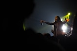 A performer with a microphone is reaching out to the audience on stage. Bright stage lights illuminate the scene from behind, creating a dynamic and energetic atmosphere. The audience is partially visible in the dark foreground, with their arms raised.