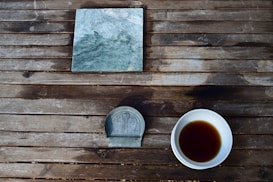 A weathered wooden table hosts a green marble tile, a small decorative metal object with a relief design, and a white bowl containing a dark liquid.