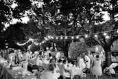 A lively outdoor gathering with people dressed in light-colored clothing sitting and standing around tables lined with decorations. The setting is under large trees with string lights creating a festive atmosphere. The scene includes adults and children, indicating a family-friendly event in a park or garden.