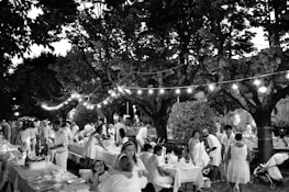 A lively outdoor gathering with people dressed in light-colored clothing sitting and standing around tables lined with decorations. The setting is under large trees with string lights creating a festive atmosphere. The scene includes adults and children, indicating a family-friendly event in a park or garden.