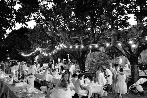 A lively outdoor gathering with people dressed in light-colored clothing sitting and standing around tables lined with decorations. The setting is under large trees with string lights creating a festive atmosphere. The scene includes adults and children, indicating a family-friendly event in a park or garden.