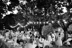 A lively outdoor gathering with people dressed in light-colored clothing sitting and standing around tables lined with decorations. The setting is under large trees with string lights creating a festive atmosphere. The scene includes adults and children, indicating a family-friendly event in a park or garden.