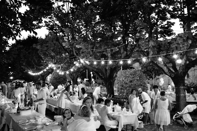 A lively outdoor gathering with people dressed in light-colored clothing sitting and standing around tables lined with decorations. The setting is under large trees with string lights creating a festive atmosphere. The scene includes adults and children, indicating a family-friendly event in a park or garden.