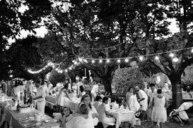 A lively outdoor gathering with people dressed in light-colored clothing sitting and standing around tables lined with decorations. The setting is under large trees with string lights creating a festive atmosphere. The scene includes adults and children, indicating a family-friendly event in a park or garden.