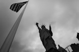 A dramatic black and white image featuring the Statue of Liberty from a low angle, to the right, alongside a tall American flag on the left. The sky is overcast with thick clouds, creating a stark backdrop.