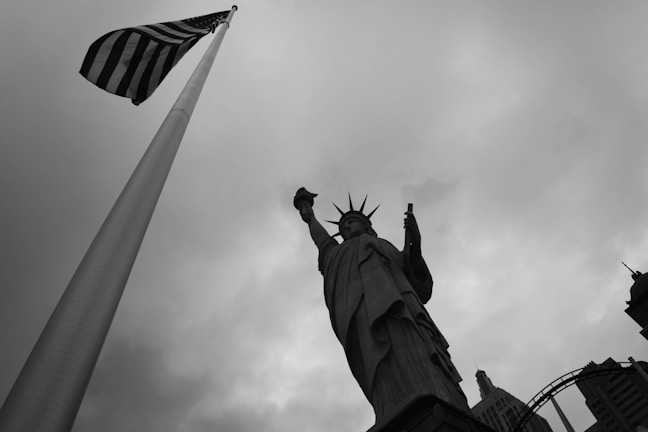 A dramatic black and white image featuring the Statue of Liberty from a low angle, to the right, alongside a tall American flag on the left. The sky is overcast with thick clouds, creating a stark backdrop.
