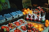 Various types of fresh fruits including strawberries, blueberries, and oranges are displayed in baskets. Candy apples wrapped in plastic and set on wooden skewers are prominently featured. The scene suggests a vibrant and bustling market setting.