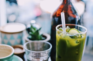 A green beverage with ice cubes and cucumber slices in a clear glass, accompanied by a straw. In the background, there is a brown glass bottle and several blurred objects, including a small succulent plant and cups.