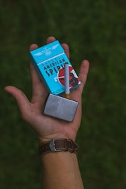 A hand holding a light blue cigarette pack labeled 'Natural American Spirit', accompanied by a single cigarette and a metallic lighter. The person wears a brown leather watch. The background is a blurred green landscape.