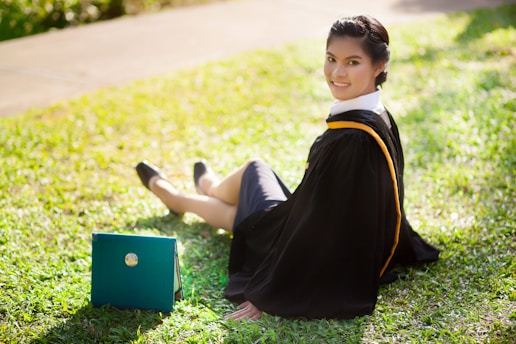 A person wearing a graduation gown is sitting on the grass while holding a diploma folder. The sunlight creates a warm and celebratory atmosphere.