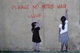 Two children are standing against a textured wall, reaching up with chalk or paint to write the words 'PLEASE NO MORE WAR' and 'LOVE' in red. The wall has a cracked appearance and the children appear to be collaborating on their anti-war message.