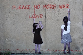 Two children are standing against a textured wall, reaching up with chalk or paint to write the words 'PLEASE NO MORE WAR' and 'LOVE' in red. The wall has a cracked appearance and the children appear to be collaborating on their anti-war message.