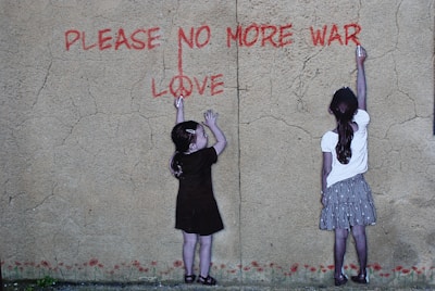Two children are standing against a textured wall, reaching up with chalk or paint to write the words 'PLEASE NO MORE WAR' and 'LOVE' in red. The wall has a cracked appearance and the children appear to be collaborating on their anti-war message.