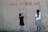 Two children are standing against a textured wall, reaching up with chalk or paint to write the words 'PLEASE NO MORE WAR' and 'LOVE' in red. The wall has a cracked appearance and the children appear to be collaborating on their anti-war message.