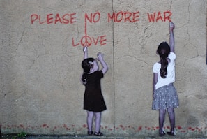 Two children are standing against a textured wall, reaching up with chalk or paint to write the words 'PLEASE NO MORE WAR' and 'LOVE' in red. The wall has a cracked appearance and the children appear to be collaborating on their anti-war message.