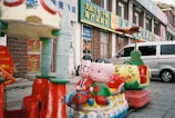A colorful children's ride featuring cartoon characters is situated on a city sidewalk. The ride includes two seats shaped like a heart with decorative elements and two large characters with smiling faces. A silver van is parked nearby, and several storefronts with Chinese signage are visible in the background.