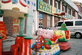 A colorful children's ride featuring cartoon characters is situated on a city sidewalk. The ride includes two seats shaped like a heart with decorative elements and two large characters with smiling faces. A silver van is parked nearby, and several storefronts with Chinese signage are visible in the background.