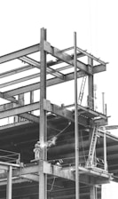 A construction site with a partially built steel framework of a structure. A welder is working on the beams, and a ladder is visible leaning against the structure. The atmosphere appears industrial and busy.
