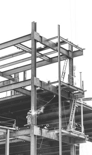 A construction site with a partially built steel framework of a structure. A welder is working on the beams, and a ladder is visible leaning against the structure. The atmosphere appears industrial and busy.