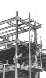 A construction site with a partially built steel framework of a structure. A welder is working on the beams, and a ladder is visible leaning against the structure. The atmosphere appears industrial and busy.