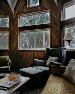 A cozy, rustic living room featuring large windows offering a view of tall trees outside. The room is furnished with a dark grey armchair with an ottoman, a vintage green chair, and a cushioned sofa with patterned pillows. There is a wooden coffee table with a magazine on top and a shaded floor lamp off to the side. The walls are adorned with wood paneling set in a herringbone pattern.