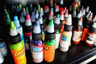 A collection of colorful ink bottles arranged closely together on a shelf. Each bottle has a twist cap, and they display a variety of bright labels with intricate designs. The inks include vibrant colors such as green, blue, red, and orange, indicating a diverse selection.