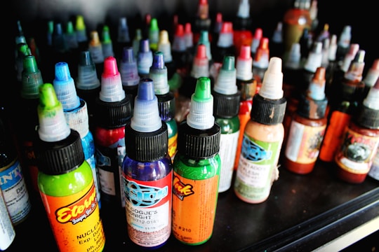 A collection of colorful ink bottles arranged closely together on a shelf. Each bottle has a twist cap, and they display a variety of bright labels with intricate designs. The inks include vibrant colors such as green, blue, red, and orange, indicating a diverse selection.