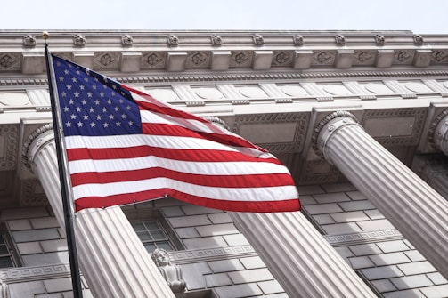 An American flag waves prominently in the foreground against a backdrop of a neoclassical building with large columns and intricate architectural details, conveying a sense of national pride.