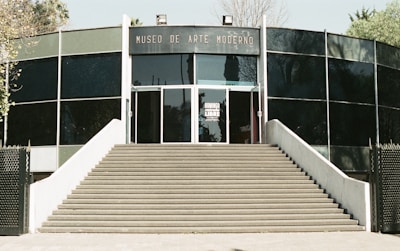 A modern building with a curved facade, featuring large glass panels and a wide staircase leading up to the entrance. The words 'Museo de Arte Moderno' are prominently displayed above the door.