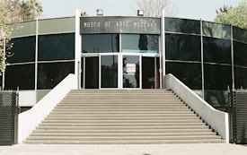 A modern building with a curved facade, featuring large glass panels and a wide staircase leading up to the entrance. The words 'Museo de Arte Moderno' are prominently displayed above the door.