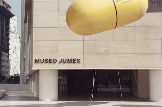 The image depicts the exterior of Museo Jumex, showcasing its modern architectural design with clean lines and a pale stone facade. A large, yellow, pill-shaped sculpture is suspended in front of the museum, attracting attention with its vibrant color. The background includes more urban architecture, suggesting a city setting.
