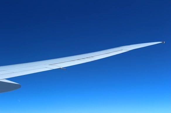 A sleek, white airplane wing extends into a clear blue sky, capturing a serene view from high altitude. The smooth gradient of the blue sky dominates the background, enhancing the clean lines of the wing.