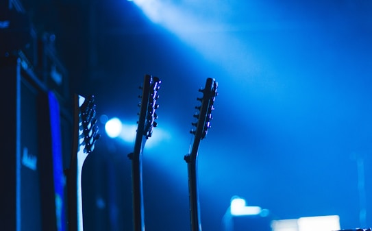 Silhouettes of multiple guitar necks and headstocks are prominently featured against a vibrant blue and black background. Amplifiers are faintly visible to the left, creating a stage-like atmosphere with dramatic lighting.