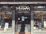 A vintage storefront for an optician, featuring weathered signage reading 'Opticians' and 'Estb 1935'. Display cases on either side showcase products like contact lenses and sunglasses, with advertisements for various eyewear brands. The building has a rustic, aged look with textured stonework surrounding the entrance.