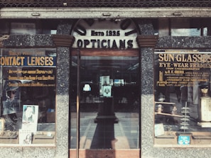 A vintage storefront for an optician, featuring weathered signage reading 'Opticians' and 'Estb 1935'. Display cases on either side showcase products like contact lenses and sunglasses, with advertisements for various eyewear brands. The building has a rustic, aged look with textured stonework surrounding the entrance.