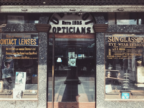 A vintage storefront for an optician, featuring weathered signage reading 'Opticians' and 'Estb 1935'. Display cases on either side showcase products like contact lenses and sunglasses, with advertisements for various eyewear brands. The building has a rustic, aged look with textured stonework surrounding the entrance.