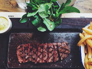A dish featuring sliced steak arranged on a dark serving platter, accompanied by a side salad with fresh greens and a portion of crispy fries. A small bowl of creamy sauce is on the side, set on a wooden table.