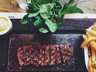A dish featuring sliced steak arranged on a dark serving platter, accompanied by a side salad with fresh greens and a portion of crispy fries. A small bowl of creamy sauce is on the side, set on a wooden table.