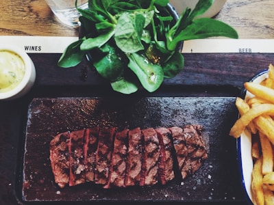A dish featuring sliced steak arranged on a dark serving platter, accompanied by a side salad with fresh greens and a portion of crispy fries. A small bowl of creamy sauce is on the side, set on a wooden table.