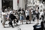 A group of people is gathered on a city street, appearing to be in movement or participating in a performance. The scene includes individuals of various ages and styles, captured in energetic postures, possibly mid-step or gesture. In the background, shop windows with mannequins are visible, suggesting an urban shopping area.