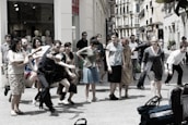 A group of people is gathered on a city street, appearing to be in movement or participating in a performance. The scene includes individuals of various ages and styles, captured in energetic postures, possibly mid-step or gesture. In the background, shop windows with mannequins are visible, suggesting an urban shopping area.