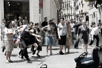 A group of people is gathered on a city street, appearing to be in movement or participating in a performance. The scene includes individuals of various ages and styles, captured in energetic postures, possibly mid-step or gesture. In the background, shop windows with mannequins are visible, suggesting an urban shopping area.