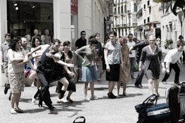 A group of people is gathered on a city street, appearing to be in movement or participating in a performance. The scene includes individuals of various ages and styles, captured in energetic postures, possibly mid-step or gesture. In the background, shop windows with mannequins are visible, suggesting an urban shopping area.