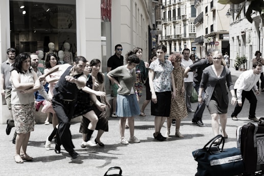 A group of people is gathered on a city street, appearing to be in movement or participating in a performance. The scene includes individuals of various ages and styles, captured in energetic postures, possibly mid-step or gesture. In the background, shop windows with mannequins are visible, suggesting an urban shopping area.