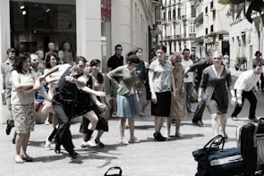 A group of people is gathered on a city street, appearing to be in movement or participating in a performance. The scene includes individuals of various ages and styles, captured in energetic postures, possibly mid-step or gesture. In the background, shop windows with mannequins are visible, suggesting an urban shopping area.