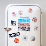 A white vintage refrigerator adorned with various colorful magnets and photos. There are images of a cityscape, a couple standing together, a heart-shaped object, a plate of food, and a circular magnet with an orange theme. The word 'FAMOUS' is spelled out using cut-up letters on separate magnets. Additional round magnets include a pattern of colorful stripes and sliced grapefruits.