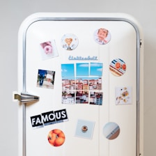 A white vintage refrigerator adorned with various colorful magnets and photos. There are images of a cityscape, a couple standing together, a heart-shaped object, a plate of food, and a circular magnet with an orange theme. The word 'FAMOUS' is spelled out using cut-up letters on separate magnets. Additional round magnets include a pattern of colorful stripes and sliced grapefruits.