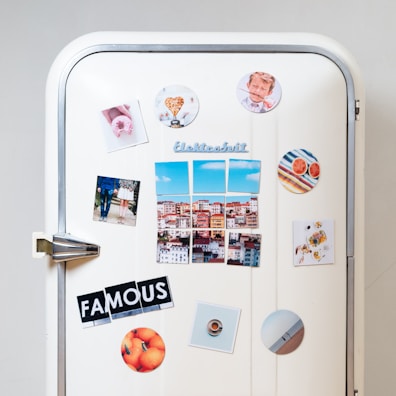 A white vintage refrigerator adorned with various colorful magnets and photos. There are images of a cityscape, a couple standing together, a heart-shaped object, a plate of food, and a circular magnet with an orange theme. The word 'FAMOUS' is spelled out using cut-up letters on separate magnets. Additional round magnets include a pattern of colorful stripes and sliced grapefruits.